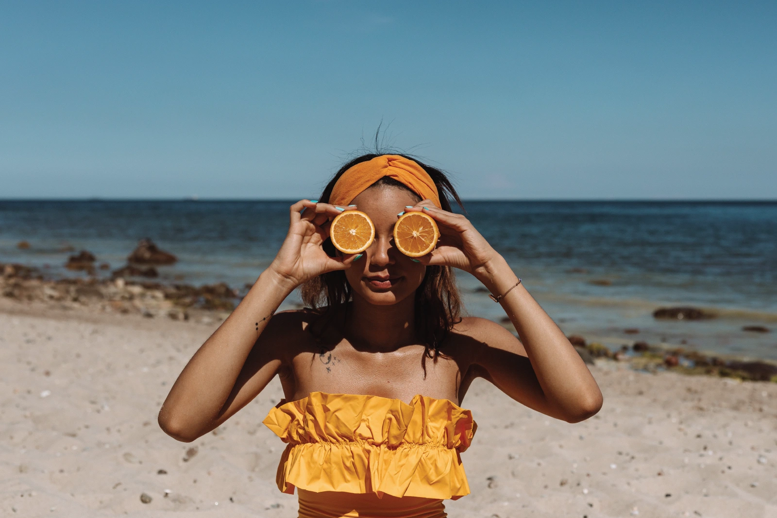 Žena na plaži drži polovice naranče ispred očiju, simbol svježine i prirodne snage ulja divlje aljaške ribe bogatog omega-3 masnim kiselinama.